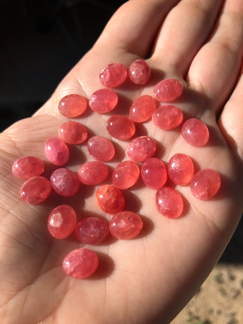 K&ouml;nnte beinhalten: Eine Sammlung von 25 ovalen, rosafarbenen und roten Edelstein-Cabochons. Die Cabochons sind glatt und poliert, mit einem leichten Schimmer.