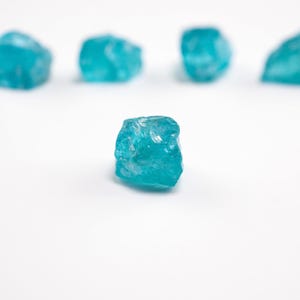 May include: A collection of five small, raw, translucent turquoise-blue mineral stones. Each stone features an irregular, jagged shape with a crystalline texture and a vibrant, saturated color. One stone is positioned in the foreground, while four others are blurred in the background.