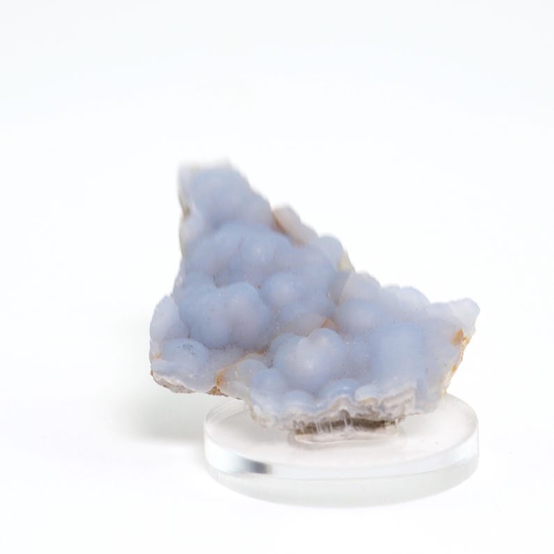 May include: A light blue chalcedony mineral specimen with a bubbly, textured surface. The stone is mounted on a clear, round display stand, showcasing its unique form and color. The mineral has a soft, cloudy appearance.