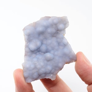 Puede incluir: Una pieza de piedra preciosa de calcedonia azul claro con una textura botrioidal y burbujeante. La piedra se sostiene entre dos dedos sobre un fondo blanco. La superficie de la piedra tiene un aspecto suave y mate.