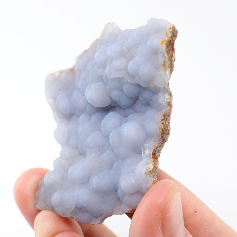 Puede incluir: Una pieza de piedra preciosa de calcedonia azul con una textura botrioidal y burbujeante. La piedra tiene un color azul claro y un borde &aacute;spero e irregular con tonos marrones y beige.