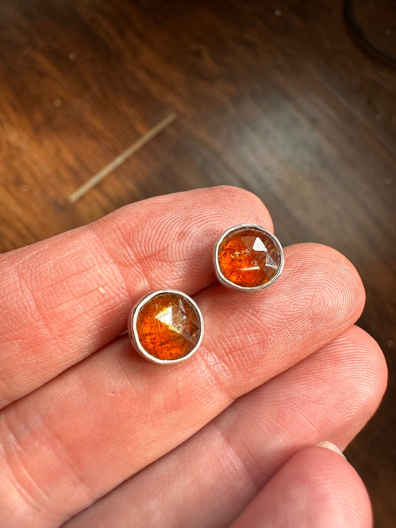 Puede incluir: Un par de pendientes de plata con detalles de piedras preciosas naranjas. Las piedras preciosas son facetadas y tienen una forma ligeramente irregular.