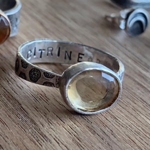 May include: A silver ring with a citrine gemstone and the word "CITRINE" engraved on the band.