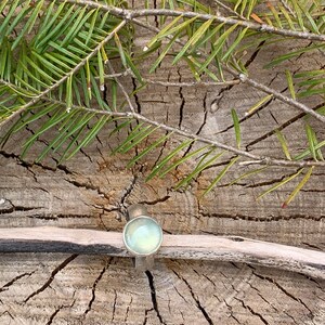 May include: A silver ring with a light green gemstone set in a round bezel. The ring is on a piece of wood with green pine needles.