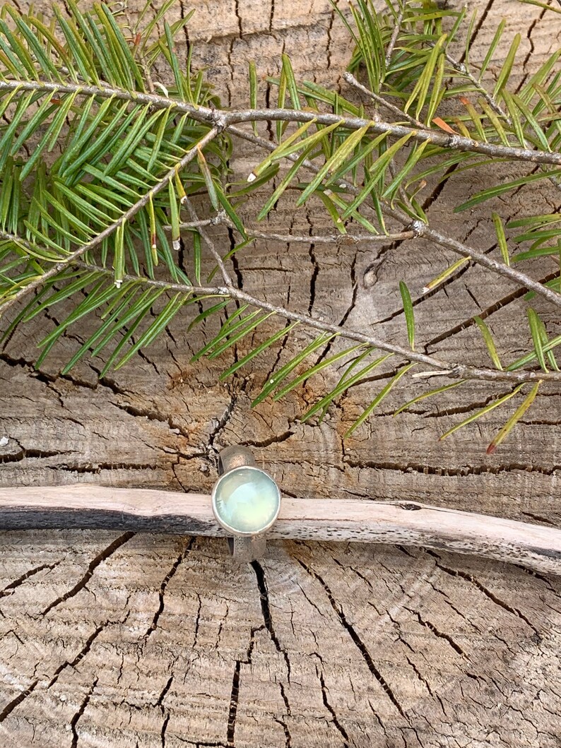 May include: A silver ring with a light green gemstone set in a round bezel. The ring is on a piece of wood with green pine needles.
