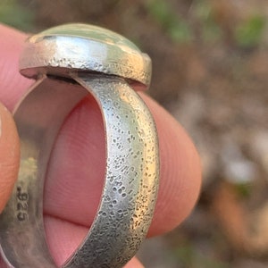 May include: A silver ring with a textured band and a smooth, oval-shaped gemstone. The ring is stamped with the number "925" on the inside of the band.