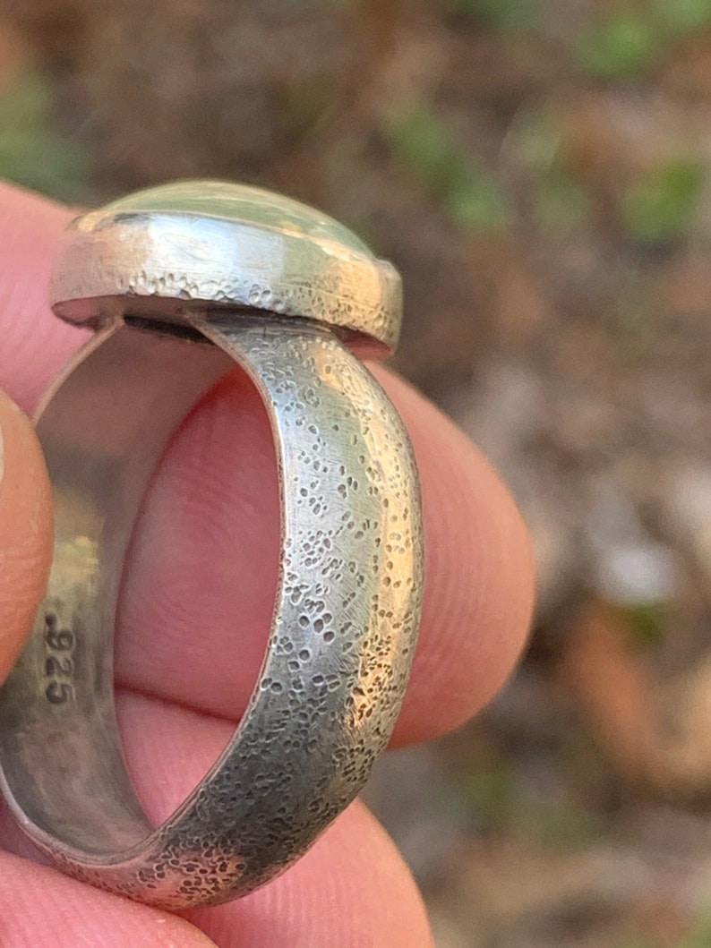 May include: A silver ring with a textured band and a smooth, oval-shaped gemstone. The ring is stamped with the number "925" on the inside of the band.