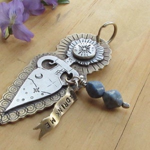 May include: A silver pendant with a detailed design of a bottle, a star, and a sunburst. The pendant is attached to a silver chain with a blue gemstone bead. The pendant has the text "Drink Me" on a banner.