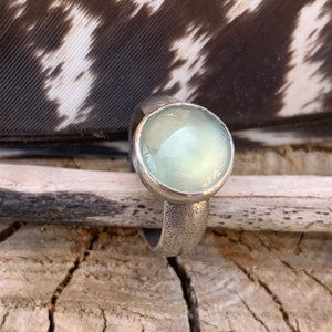 May include: A silver ring with a light green gemstone set in a bezel setting. The ring is textured and has a hammered finish.