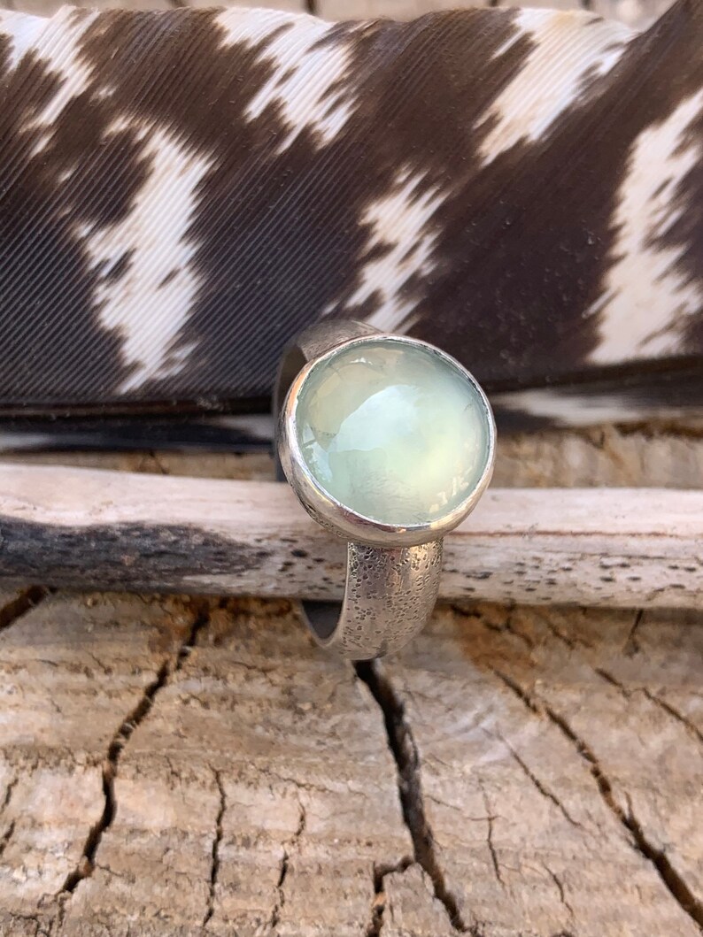 May include: A silver ring with a light green gemstone set in a bezel setting. The ring is textured and has a hammered finish.