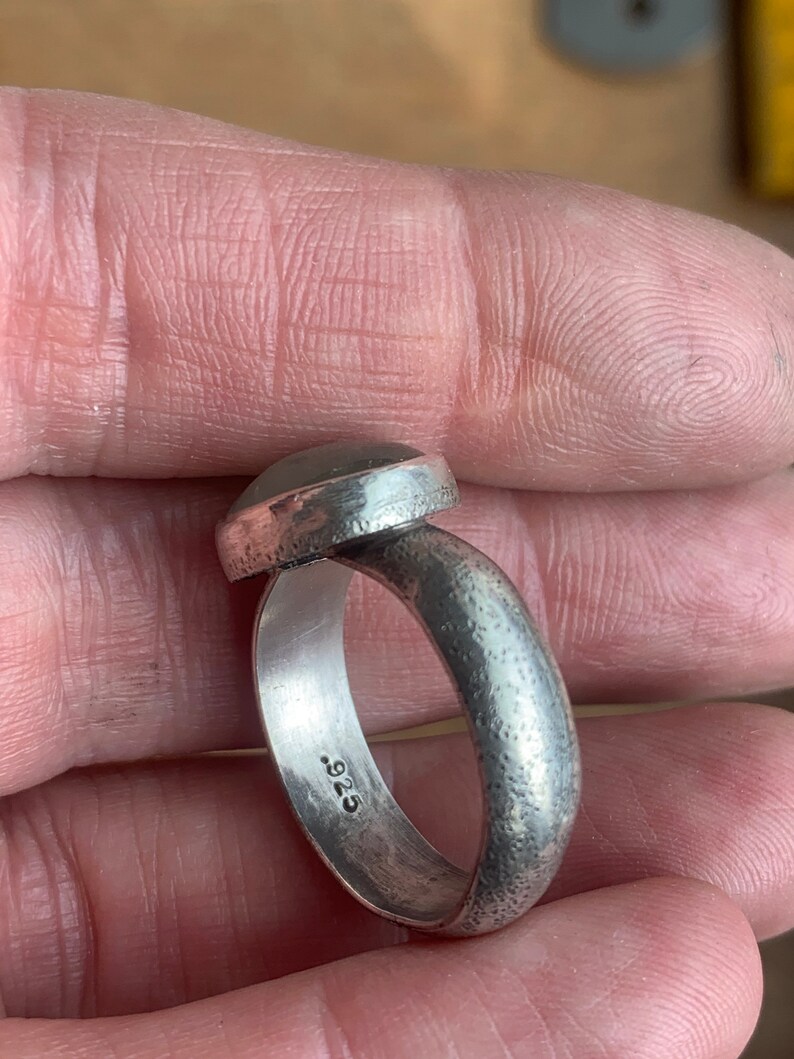 May include: A silver ring with a textured band and a round, smooth gemstone. The ring is stamped with the number "925" on the inside of the band.