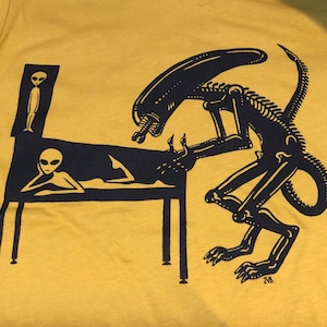 May include: A yellow t-shirt with a black and white graphic print of an alien creature standing over a reclining alien. The alien creature has a long tail and is holding a weapon. The text "JVB" is printed in the bottom right corner of the graphic.