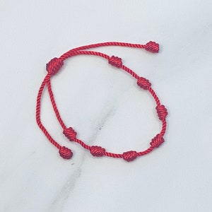 Puede incluir: Una pulsera de cuerda roja con múltiples nudos. La pulsera está hecha de un cordón fino y flexible y presenta varios nudos espaciados a lo largo de su longitud. Los extremos de la pulsera tienen nudos ajustables para el tamaño.