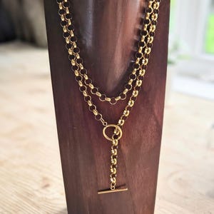 May include: A gold-coloured necklace with a toggle clasp, displayed on a wooden jewellery stand. The necklace features multiple strands of linked chain, with a circular pendant and a bar closure. The jewellery stand is a dark brown colour.