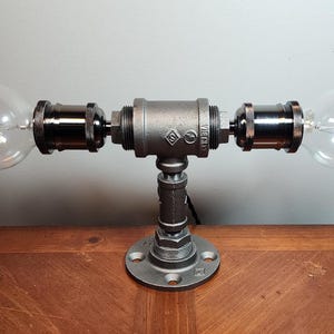 May include: A gray metal table lamp with two clear glass light bulbs and a black metal base. The lamp is on a wooden surface.