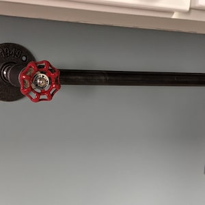 May include: A black metal pipe towel rack with a red valve handle. The pipe is attached to a wall with a black metal flange. The flange has the text "1949" on it. The rack is mounted on a light blue wall.