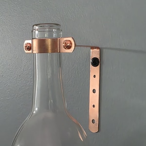 May include: A clear glass bottle with a copper-colored metal bracket attached. The bracket is designed to hold the bottle, with screws securing it. The bottle is partially visible, showcasing its shape and the bracket's design.