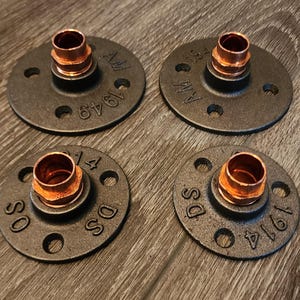 May include: Four industrial-style pipe flanges with copper fittings. The flanges are dark gray with circular holes and stamped text. The copper fittings are a warm orange color. The items are arranged on a wood-grain surface.