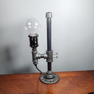 May include: A black metal table lamp with a clear glass globe light bulb. The lamp is made of industrial pipe fittings and has a black metal base.