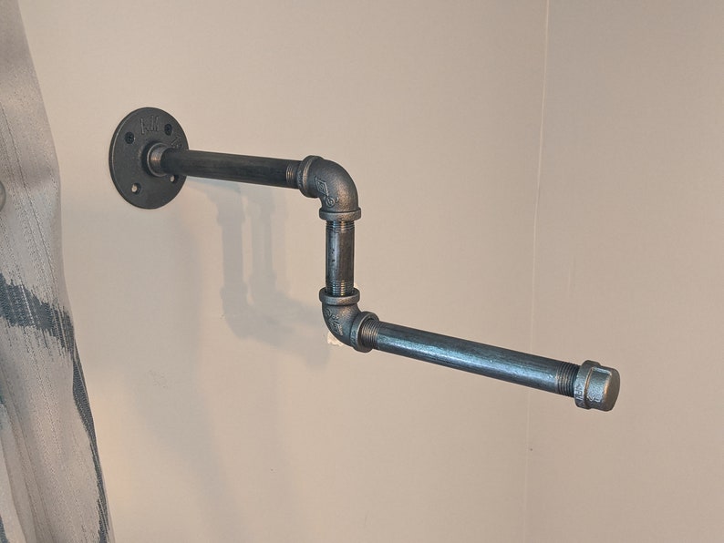 K&ouml;nnte beinhalten: Ein schwarzer Metall-Handtuchhalter, der an einer Wand montiert ist. Der Halter hat eine Flansch an der Wand und ein horizontales Rohr, das von ihm wegf&uuml;hrt. Das Rohr ist in einem 90-Grad-Winkel gebogen und erstreckt sich dann wieder horizontal.