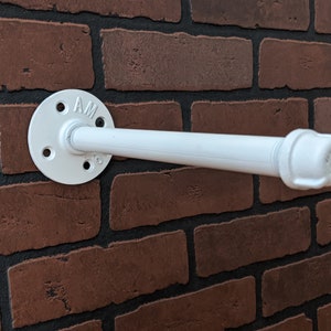 May include: White metal pipe shelf bracket with a flange and a cap. The bracket is mounted to a brick wall.