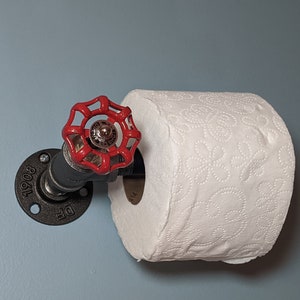 May include: A black metal toilet paper holder with a red valve handle. The holder is mounted to a wall and has a roll of white toilet paper on it.