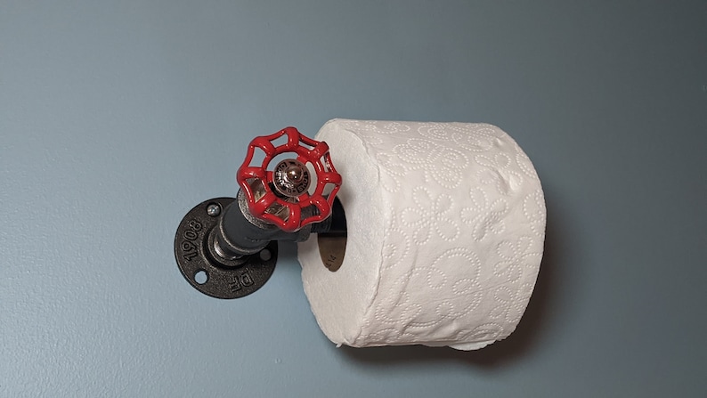 May include: A black metal toilet paper holder with a red valve handle. The holder is mounted to a wall and has a roll of white toilet paper on it.