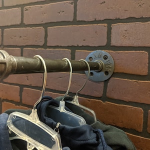 May include: A metal pipe with a flange attached to a brick wall. The pipe has three clear plastic hangers hanging from it. The flange has the text "1949" on it.