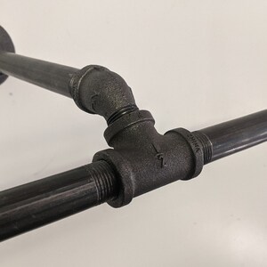 K&ouml;nnte beinhalten: Schwarze Metallrohrverbindungen bilden eine T-f&ouml;rmige Regalhalterung. Die Halterung ist mit einer schwarzen Metallflansch an einer Wand befestigt.