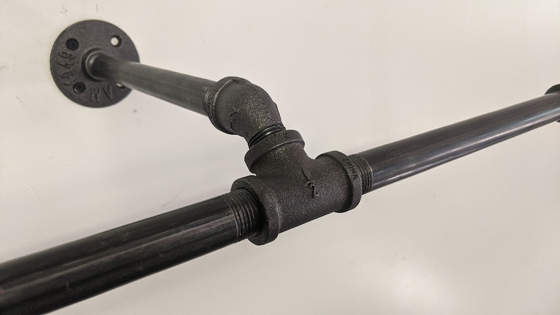 K&ouml;nnte beinhalten: Schwarze Metallrohrverbindungen bilden eine T-f&ouml;rmige Regalhalterung. Die Halterung ist mit einer schwarzen Metallflansch an einer Wand befestigt.