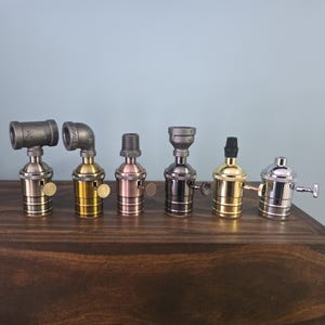 May include: A collection of six different lamp holder sockets in various metallic finishes, including brass, copper, and silver. The sockets have different shapes and styles, with some featuring pipe fittings. The background is a light blue wall.