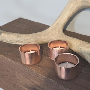 May include: Three copper candle holders with white candles on a wooden surface. The candle holders are round and have a smooth, polished finish.