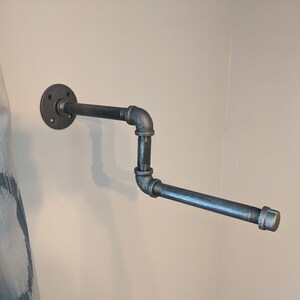 K&ouml;nnte beinhalten: Ein schwarzer Toilettenpapierhalter aus Metall, der an einer wei&szlig;en Wand montiert ist. Der Halter besteht aus industriellen Rohrverbindungsst&uuml;cken und hat ein schlichtes, modernes Design.