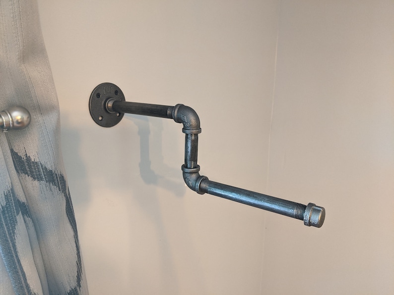 K&ouml;nnte beinhalten: Ein schwarzer Toilettenpapierhalter aus Metall, der an einer wei&szlig;en Wand montiert ist. Der Halter besteht aus industriellen Rohrverbindungsst&uuml;cken und hat ein schlichtes, modernes Design.