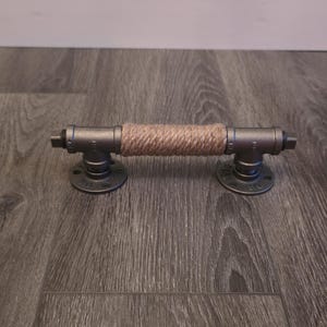 May include: Industrial-style towel bar made from galvanized steel pipes and fittings. The center section is wrapped with natural-colored rope. The bar has a dark gray finish and is mounted on circular flanges for secure installation.
