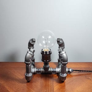 May include: A gray metal table lamp with a clear glass light bulb. The lamp is made of industrial pipe fittings and has a black cord.