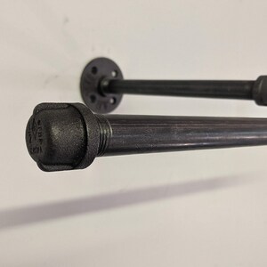 K&ouml;nnte beinhalten: Schwarzes Metallrohrregal mit einer Flansch an der Wand und einem schwarzen Rohrbogenverbinder. Das Regal besteht aus industriellen Rohrverbindungsst&uuml;cken.