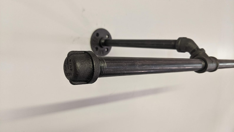 K&ouml;nnte beinhalten: Schwarzes Metallrohrregal mit einer Flansch an der Wand und einem schwarzen Rohrbogenverbinder. Das Regal besteht aus industriellen Rohrverbindungsst&uuml;cken.