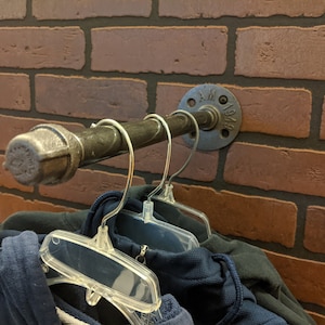 May include: A black metal pipe with a flange and a black pipe cap is mounted to a brick wall. Two clear plastic hangers are hanging from the pipe. Two navy blue hoodies are hanging on the hangers.