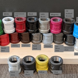 May include: A variety of colored metal pipe fittings, including brass, green, white, red, black, satin nickel, oil rubbed bronze, magenta, oasis blue, Venetian bronze, navy blue, ink blue, apple red, cherry, espresso, dark steel, primed, gun metal gray, hammered black, ford gray, flat black, yellow, and clear. The fittings are arranged in rows on a wooden surface.