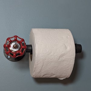 May include: A red industrial-style toilet paper holder with a black pipe and a red valve handle. The holder is mounted on a light blue wall.