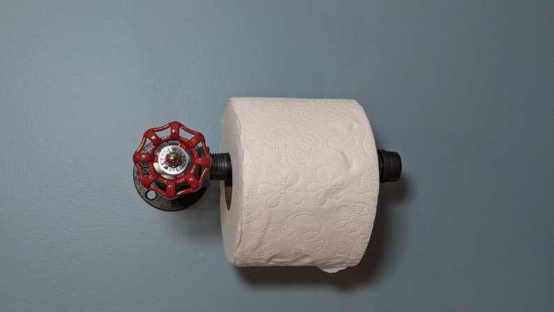May include: A red industrial-style toilet paper holder with a black pipe and a red valve handle. The holder is mounted on a light blue wall.
