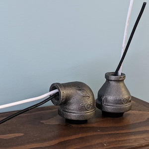 May include: Two industrial-style metal pipe fittings, one elbow and one vase-shaped, each holding a reed diffuser stick. The elbow fitting holds a white and a black stick, while the vase holds a black stick. The fittings are on a wooden surface.
