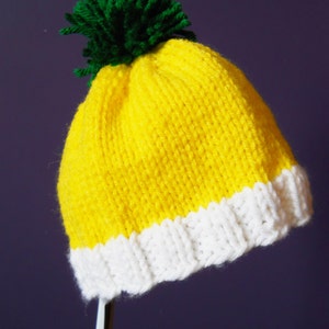 May include: A yellow knitted hat with a white brim and a green pom-pom on top.