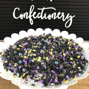 May include: A white bowl filled with black, yellow, and purple Halloween sprinkles. The sprinkles are shaped like bats, stars, and round beads. The bowl is sitting on a burlap surface.