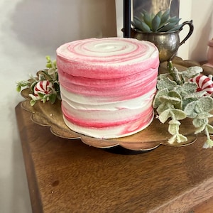 May include: A round, layered cake with pink and white frosting, displayed on a gold-colored, scalloped-edge tray. The cake is decorated with a swirl pattern on top. The tray sits on a wooden surface, with decorative greenery and peppermint candies around the base.