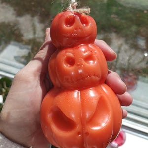 May include: A stack of three orange wax pumpkins shaped into a vertical figure with carved facial expressions. The top pumpkin features a small white wick tied with a piece of natural twine. The surface of the wax has a subtle glittery finish.
