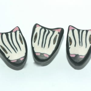 May include: Three handmade polymer clay zebra-themed pendants. Each pendant is black with white stripes, pink accents, and a pink nose. The pendants are arranged on a white background.