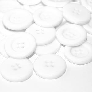 May include: A collection of white plastic buttons with four holes each. The buttons are arranged in a random pattern on a white surface.