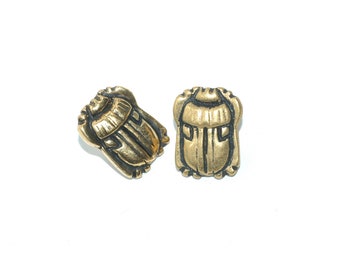May include: Two antique gold-coloured scarab beetle charms. The charms have etched lines and a textured surface, set against a white background. Suitable for jewellery making or other craft projects, these charms are detailed.